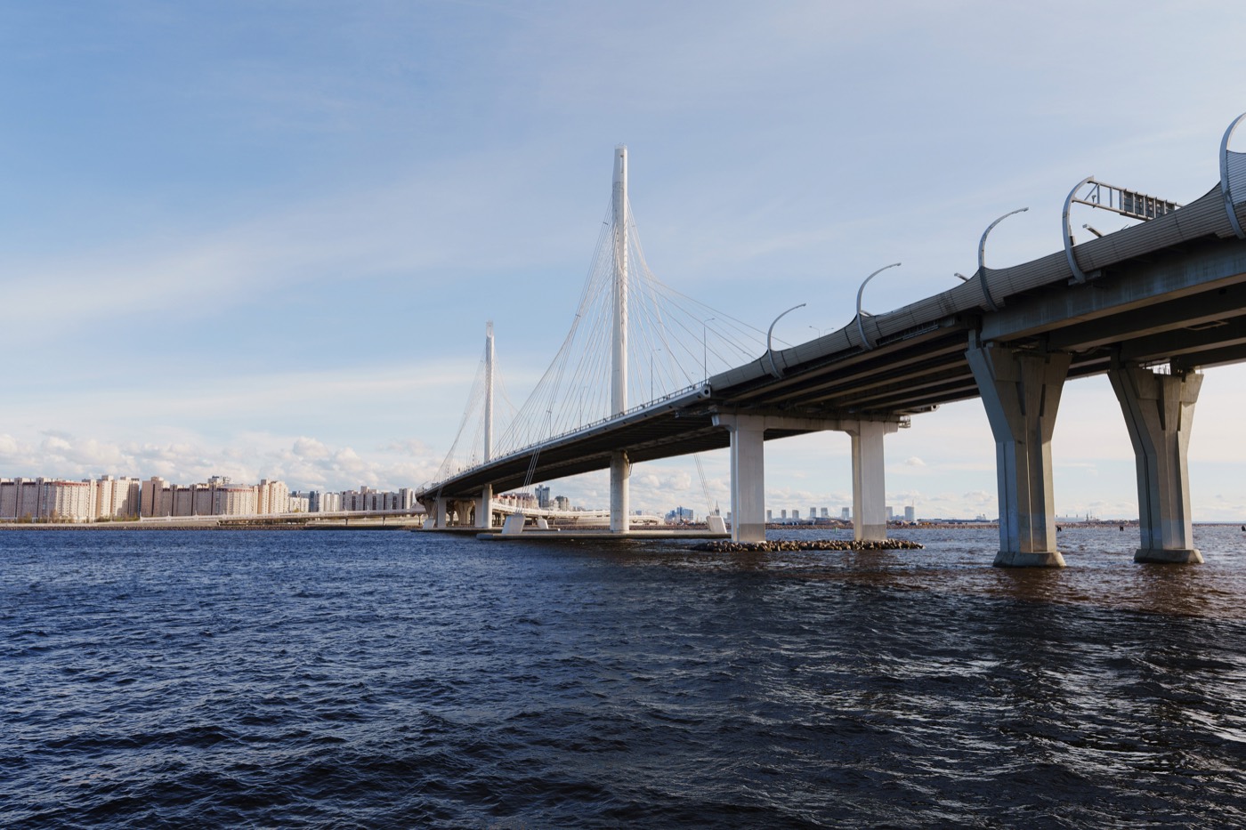 Вантовый мост над водой: широкий пролёт на опорах уходит к берегу с домами, серо-голубое небо
